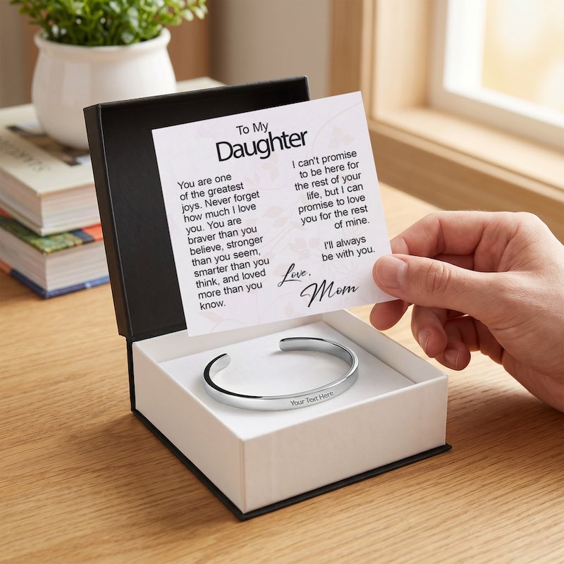 Close-up lifestyle shot of a hand revealing a printed "To My Daughter" message card signed "Love, Mom," displayed inside a gift box above a custom silver cuff bracelet with "Your Text Here" sample engraving.