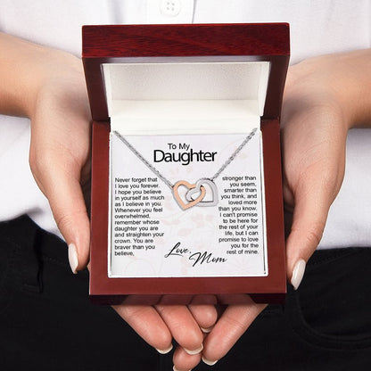 A person holds an open dark wood gift box containing a silver necklace with two interlocking heart shapes on a printed message card addressed 'To My Daughter.'.