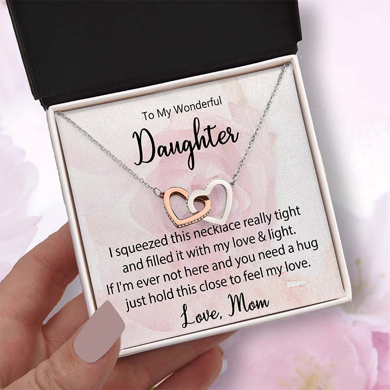 Two-tone interlocking heart necklace displayed in a gift box with a heartfelt daughter message card from Mom, held in hand against a soft pink floral background