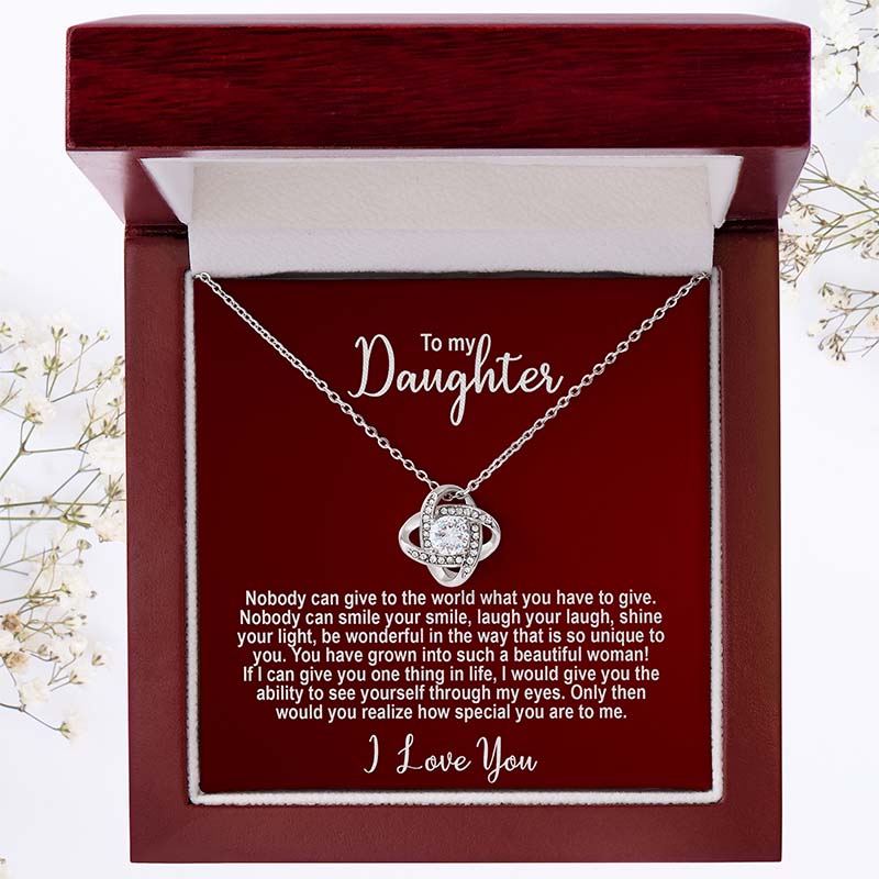 A red card with a personalized message for a daughter accompanies a silver knot necklace, both stored in a wooden box.