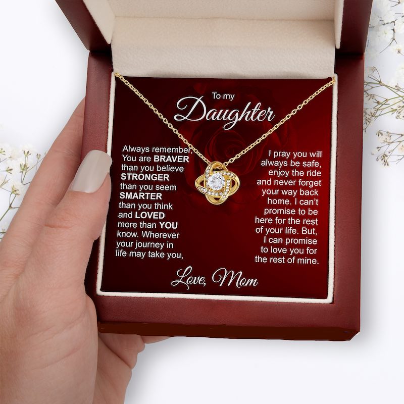 A gold knot necklace with a clear central stone is displayed inside a red gift box on a printed message card addressed 'To my Daughter.' A hand holds the box open.