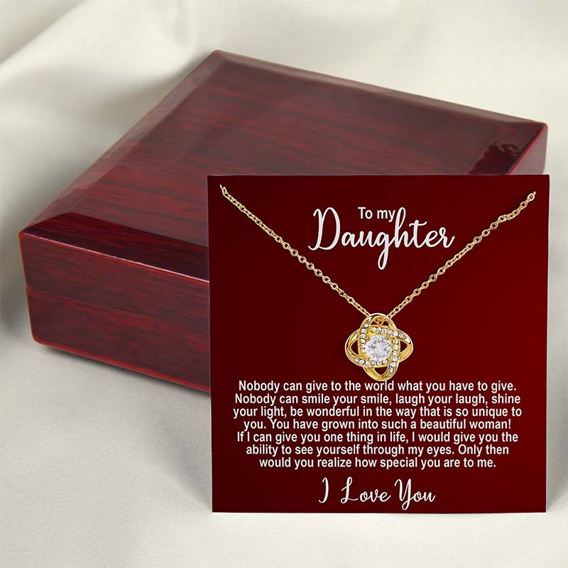 Gold knot necklace with a sentimental message card for a daughter on a red card displayed on a red wooden box against a light silk background.