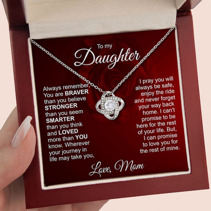 A silver necklace with a heart-shaped pendant set with small clear stones, presented inside a red gift box with a message card addressed 'To my Daughter'. A hand with manicured nails is holding the box on the left.