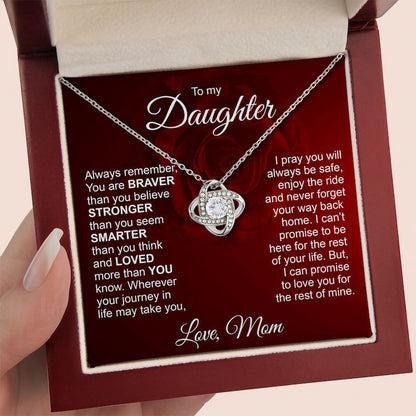 A silver necklace with a heart-shaped pendant set with small clear stones, presented inside a red gift box with a message card addressed 'To my Daughter'. A hand with manicured nails is holding the box on the left.
