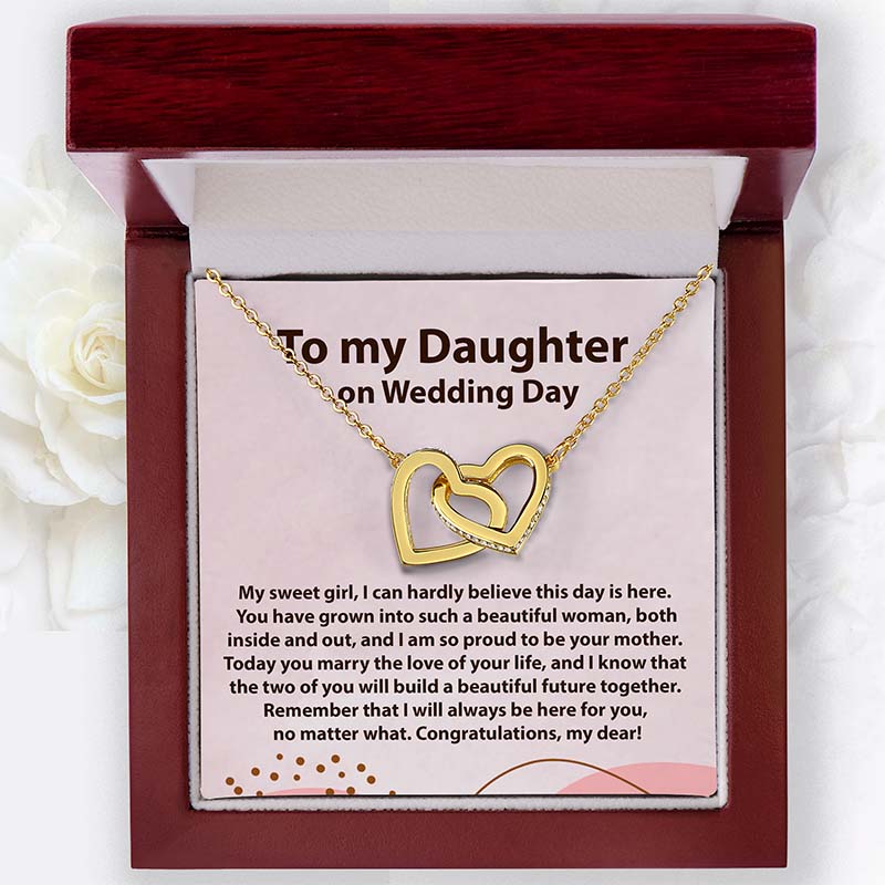 A gold double-heart pendant necklace presented inside an open dark wood gift box with a white insert card that reads 'To my Daughter on Wedding Day' and a congratulatory message. White flowers are softly blurred in the background.