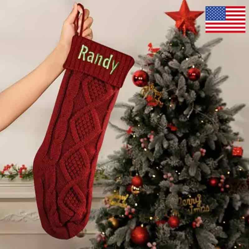 Red cable-knit Christmas stocking with green embroidered name Randy held up beside a decorated Christmas tree with red ornaments.   