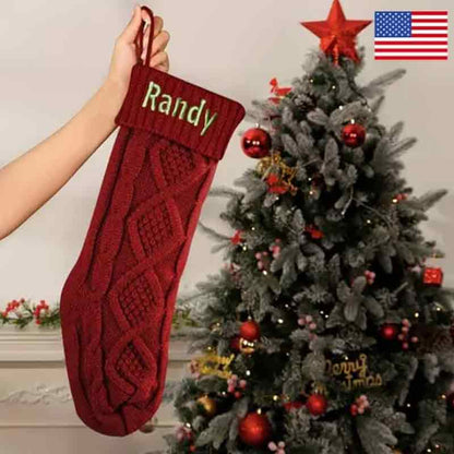 Red cable-knit Christmas stocking with green embroidered name Randy held up beside a decorated Christmas tree with red ornaments.   