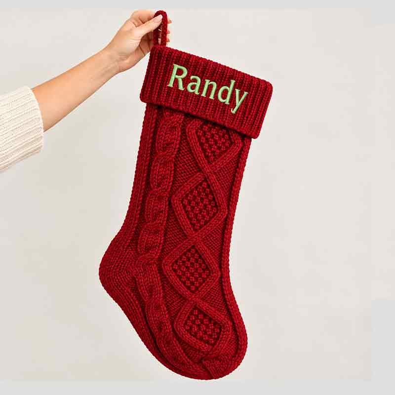 Hand holding a deep red cable-knit Christmas stocking against a plain white background, featuring the name Randy embroidered in green on the folded cuff and intricate diamond knit patterns.
