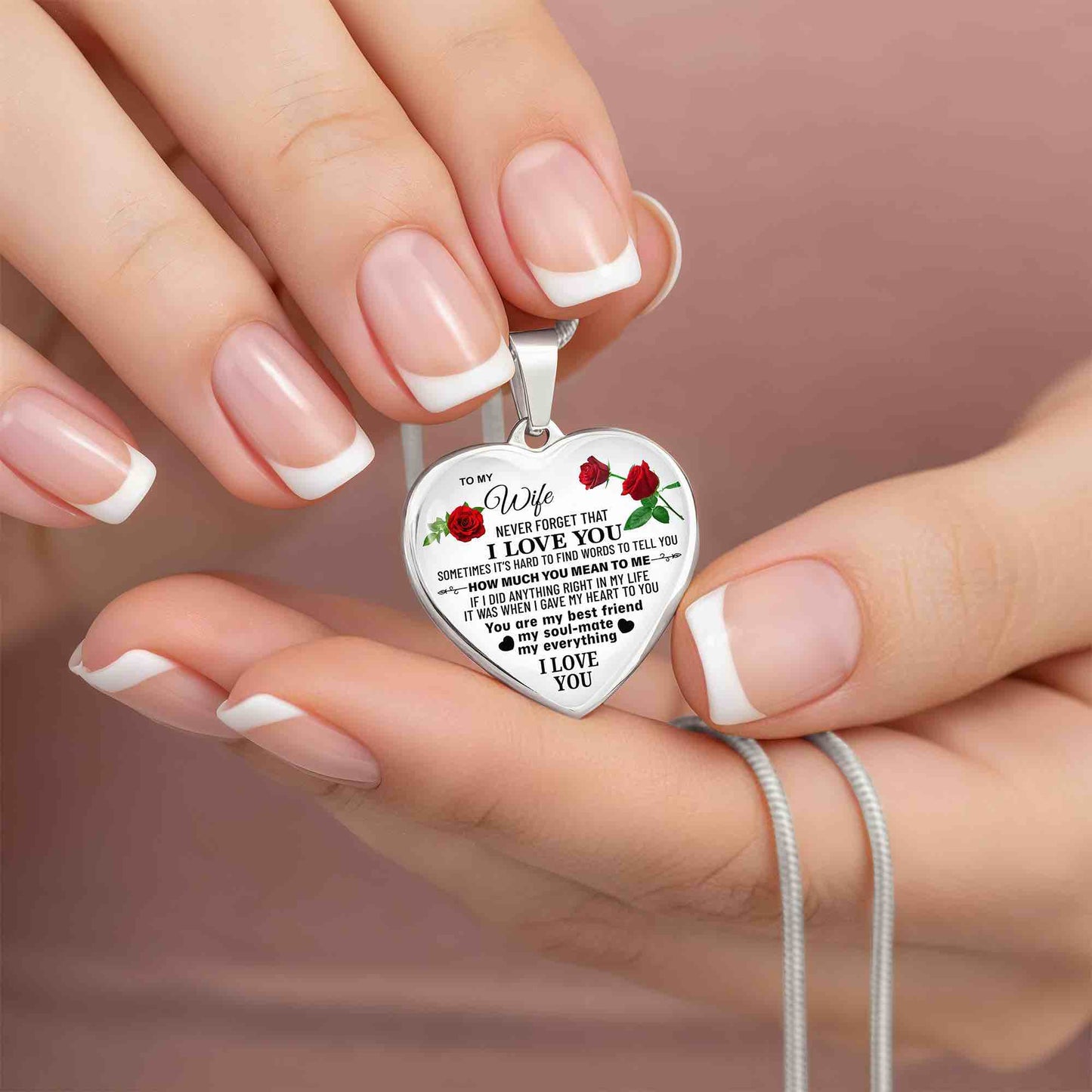 Engraved silver heart necklace with a romantic message for wife and red rose accents on a silver snake chain, shown in close-up, held by hands.