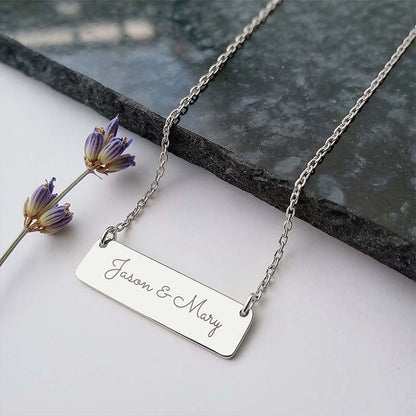 A silver rectangular nameplate necklace engraved with 'Jason & Mary' in cursive script, displayed on a white surface with a dark gray stone slab and dried lavender flowers as props.