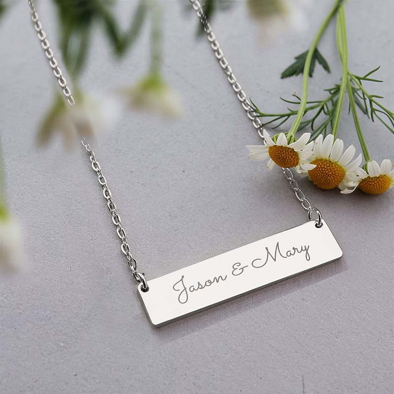 A silver rectangular bar necklace engraved with the names 'Jason & Mary' in cursive, displayed on a gray surface with white daisies in the background.