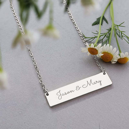 A silver rectangular bar necklace engraved with the names 'Jason & Mary' in cursive, displayed on a gray surface with white daisies in the background.
