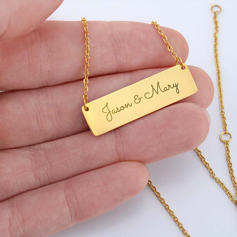 A gold rectangular nameplate necklace engraved with 'Jason & Mary' in script, held in a person's hand against a white background.