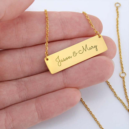 A gold rectangular nameplate necklace engraved with 'Jason & Mary' in script, held in a person's hand against a white background.