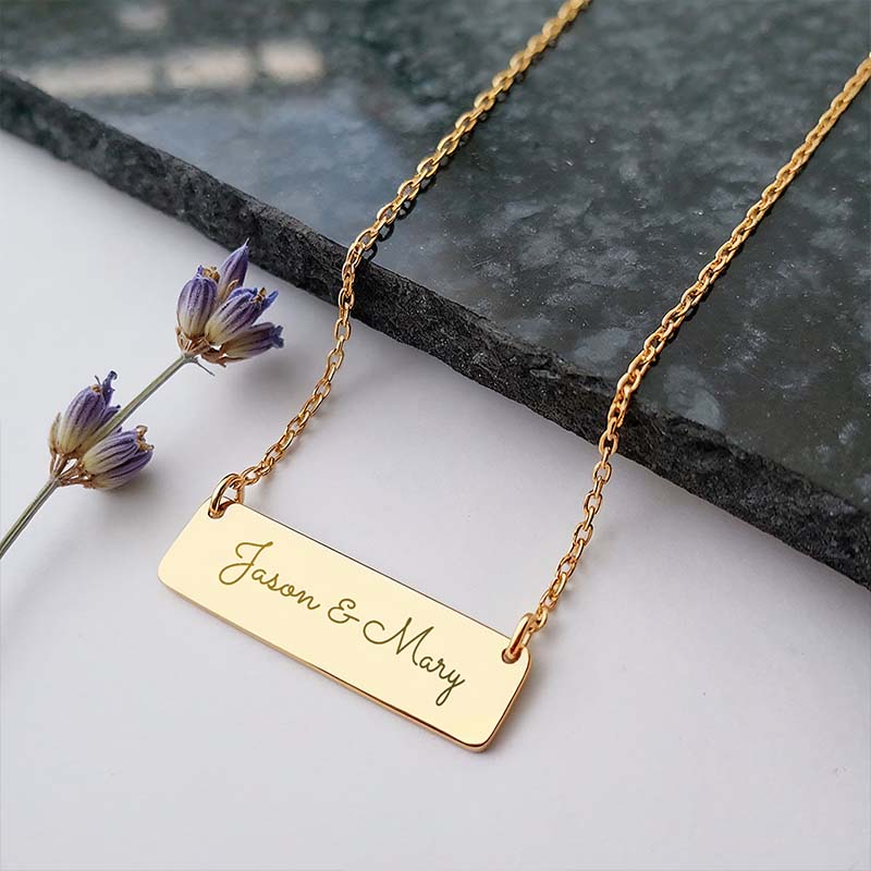Gold necklace with 'Jason & Mary' engraving bar pendant on a stone surface with lavender flowers.