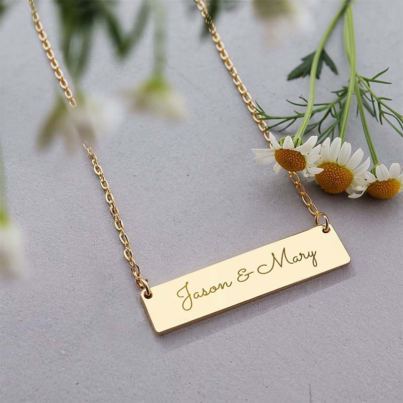 A gold bar necklace with the names 'Jason & Mary' engraved in cursive, styled with white daisies on a textured gray background.