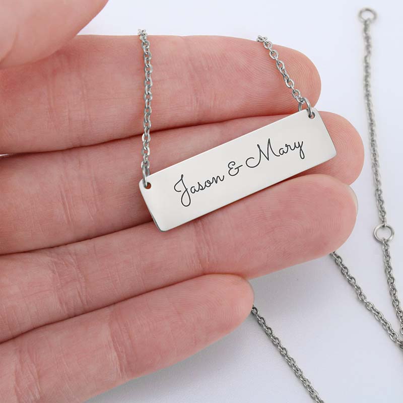 A silver rectangular bar necklace engraved with 'Jason & Mary' in black script, held in a person's hand against a white background.