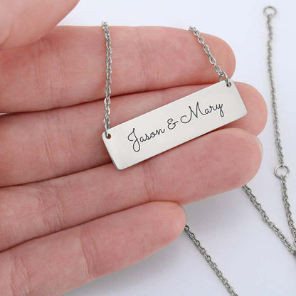 A silver rectangular bar necklace engraved with 'Jason & Mary' in black script, held in a person's hand against a white background.