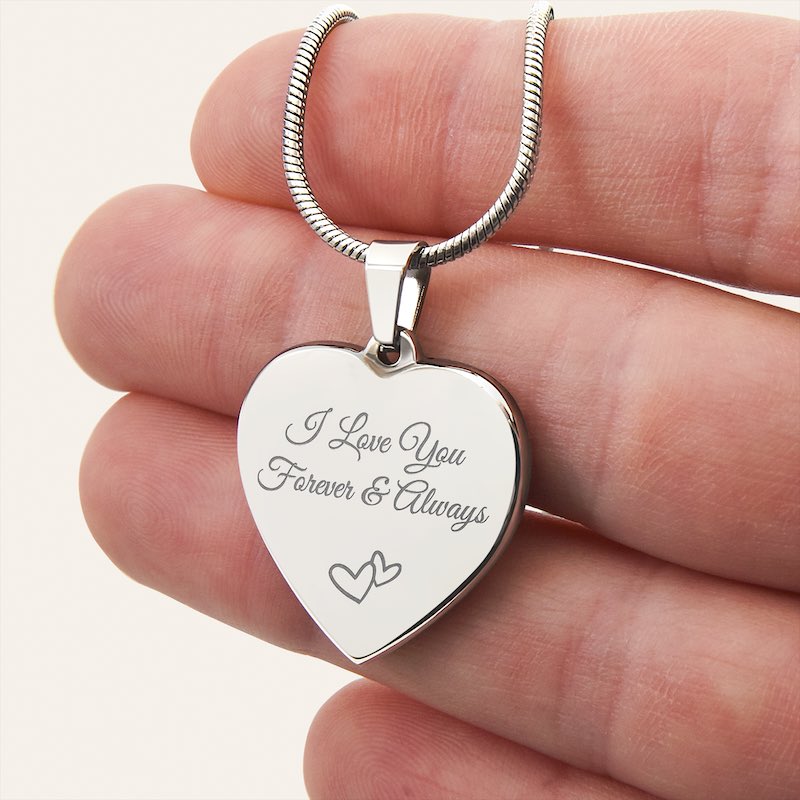 Heart-shaped silver pendant with 'I Love You Forever & Always' engraving held by hand fingers.