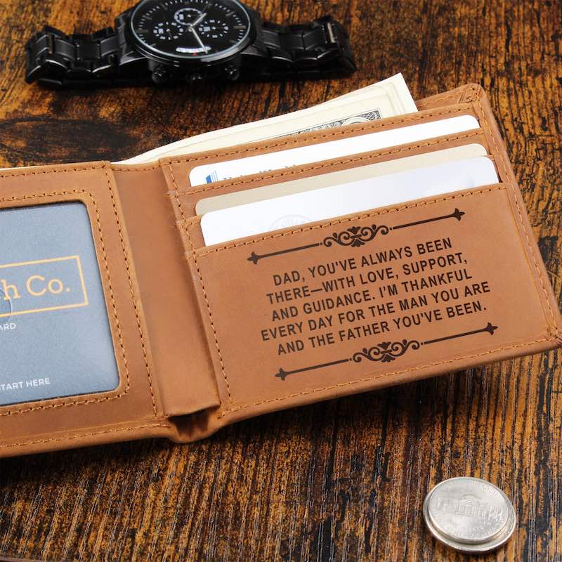 Open tan leather bifold wallet resting on a rustic dark wood surface with paper currency tucked inside, flanked by a black metal watch above and a silver coin below, featuring card slots engraved with a message of love and gratitude for Dad.   