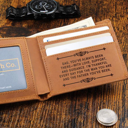Open tan leather bifold wallet resting on a rustic dark wood surface with paper currency tucked inside, flanked by a black metal watch above and a silver coin below, featuring card slots engraved with a message of love and gratitude for Dad.   