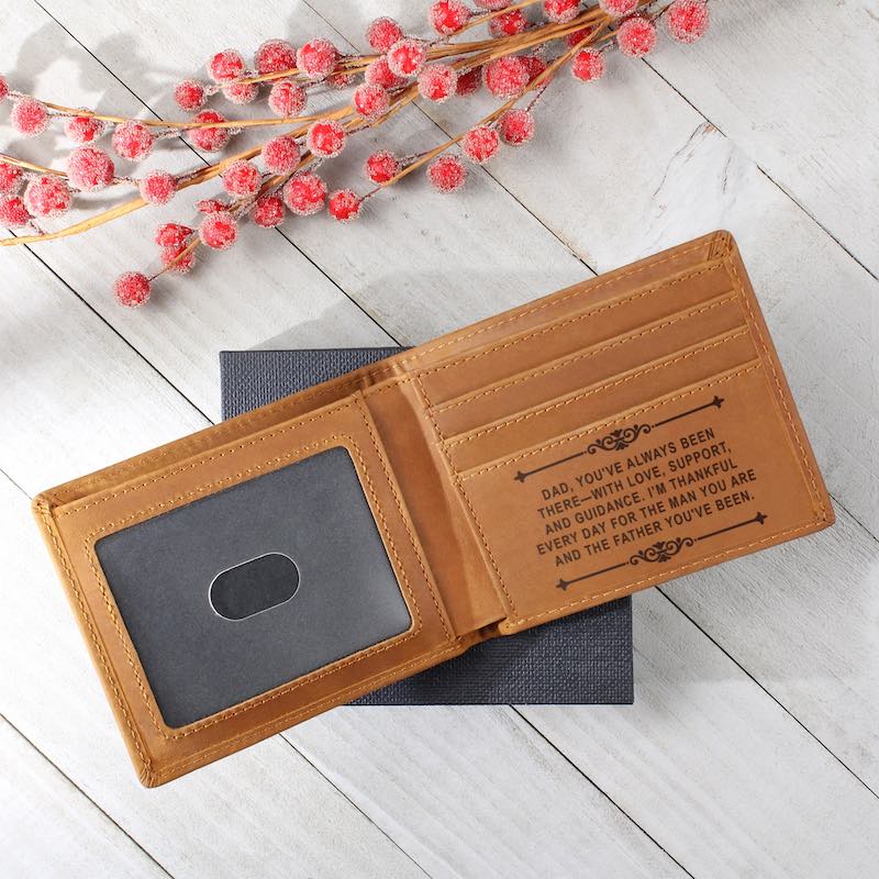 Tan bifold leather wallet resting on a dark gift box against a whitewashed wooden background with frosted red berries, showcasing card slots laser-engraved with a sentimental message for Dad.   