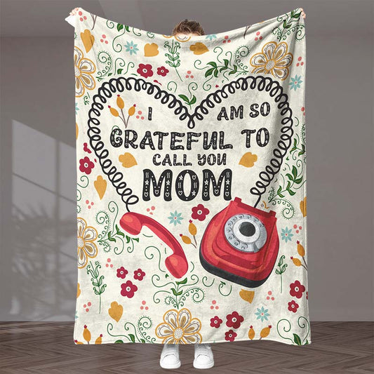 Blanket with 'I am so grateful to call you Mom' text and floral design, with a heart outline and red vintage telephone illustration, held by a person in an indoor background.