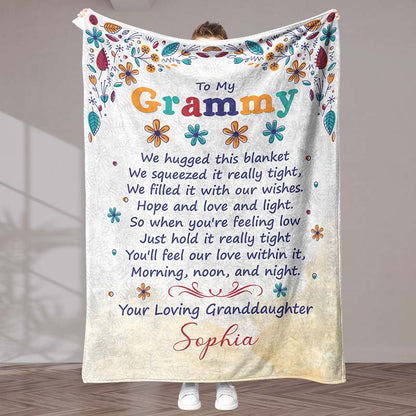 Beige blanket with floral design and sentimental text 'To My Grammy', held by a person, in an indoor background.