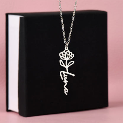 Silver flower birth name necklace and November chrysanthemum pendant in front of a black gift box with white edges, set against a soft pink background