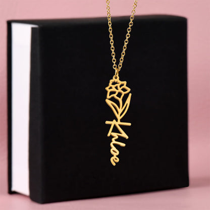 Flower birth gold necklace with a December narcissus flower and scripted name pendant on a black and white box against a pink background