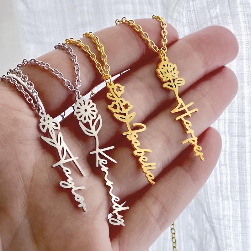 A hand holding dainty gold and silver necklaces with flower designs and personalized name pendants.