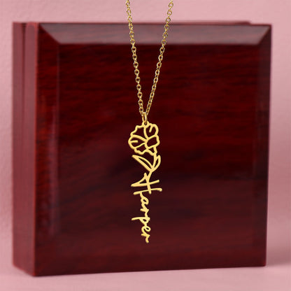 Gold flower birth name necklace in front of a mahogany gift box set against a soft pink background