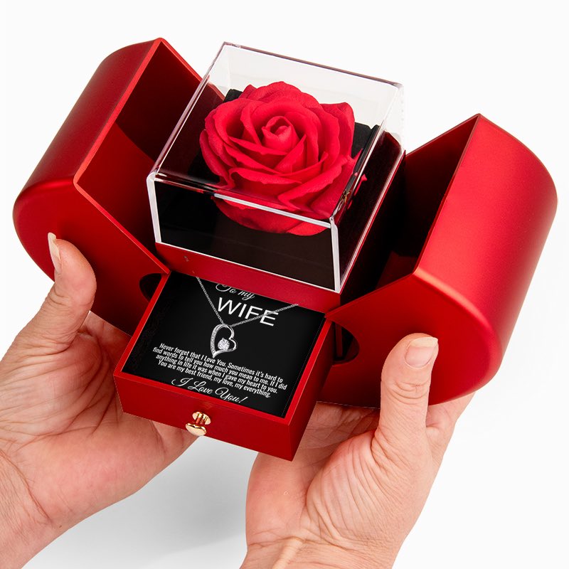 A red rose preserved in a clear square display box, presented inside an open red gift box with a pull-out message card addressed to 'My Wife.' Hands are holding the gift box against a clean white background.