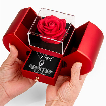 A red rose preserved in a clear square display box, presented inside an open red gift box with a pull-out message card addressed to 'My Wife.' Hands are holding the gift box against a clean white background.