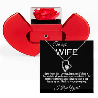 A red heart-shaped jewelry gift box with a clear lid showing a red rose, alongside a black gift card addressed to a wife with a silver heart pendant necklace and a sentimental message.