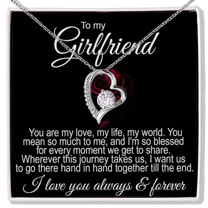 Gifts for Girlfriend: A silver heart-shaped necklace featuring a round sparkling crystal at its center, presented inside a black gift box. Above and below the necklace is a printed romantic message addressed "To my Girlfriend," expressing love and commitment.   