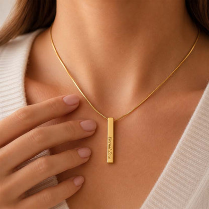 Gold necklace with a vertical bar pendant engraved on a person wearing a light-colored sweater.