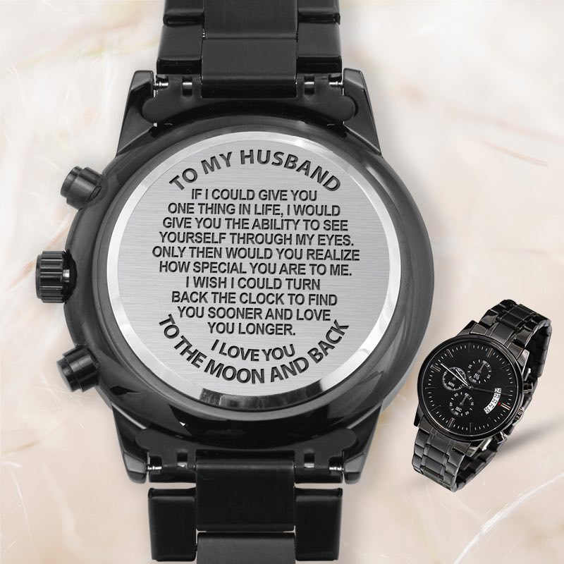 Black stainless steel watch shown from the back with engraved sentimental text for a husband, alongside a smaller front view of the black chronograph watch on a light marble background.