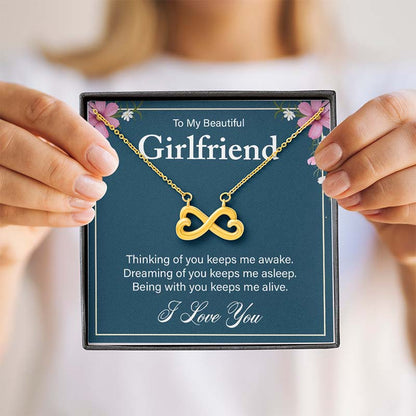 Hands holding a gift box containing a gold infinity heart necklace on a blue card reading "To My Beautiful Girlfriend