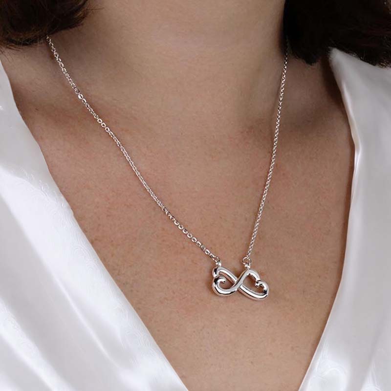 A close-up of a silver infinity heart pendant necklace worn by a model in a white satin top.