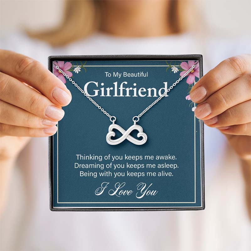 Hands holding a gift box containing a silver infinity heart necklace on a blue card reading "To My Beautiful Girlfriend