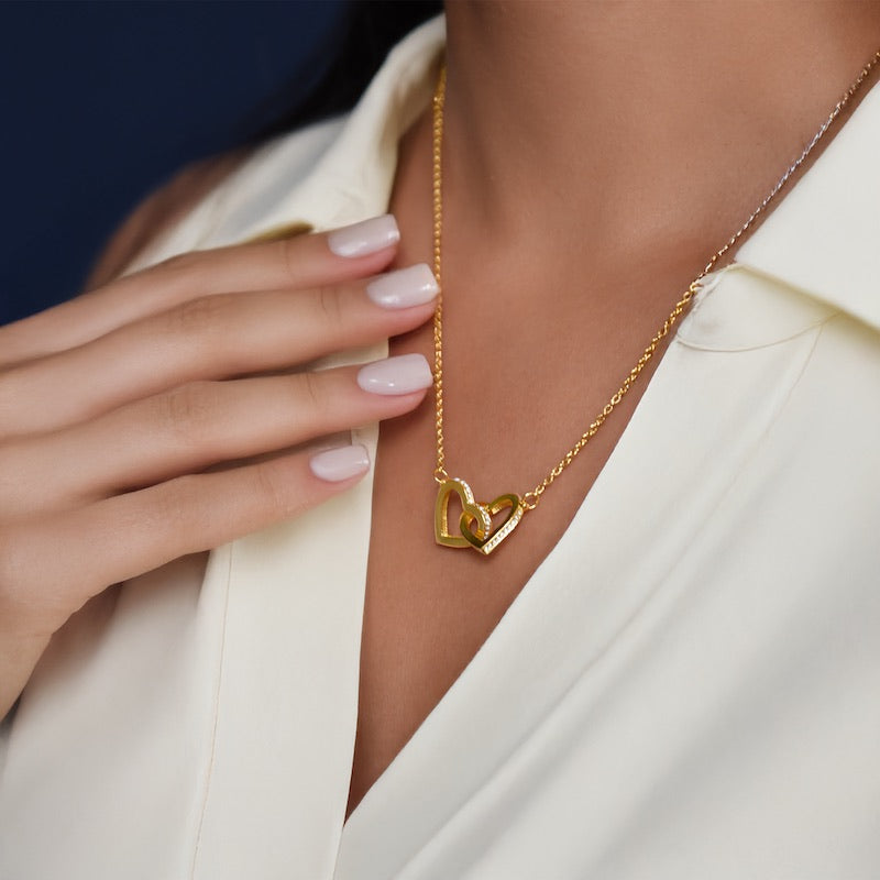A model wearing a gold necklace featuring two interlocking heart-shaped pendants studded with small crystals, paired with a off-white collared shirt.