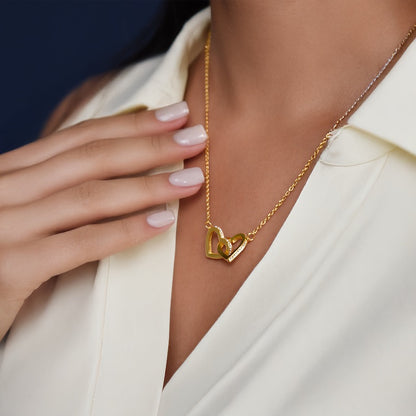 A model wearing a gold necklace featuring two interlocking heart-shaped pendants studded with small crystals, paired with a off-white collared shirt.