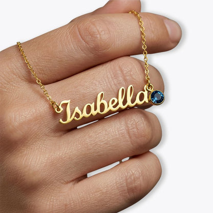 A gold nameplate necklace spelling 'Isabella' worn on a person's hand, featuring a small round blue gemstone accent at the top right of the pendant, set against a clean white background.