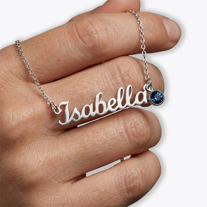 A close-up of a hand wearing a silver nameplate necklace that reads 'Isabella', with a small round blue gemstone charm on the chain, against a light gray background.