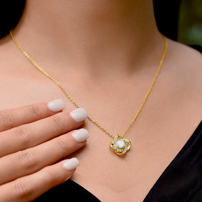 A close-up of a person wearing a delicate gold chain necklace with a small gold pendant featuring a white center stone and clear surrounding stones. The person's hand with light pink manicured nails rests near the pendant, and they are wearing a black top against a dark background.