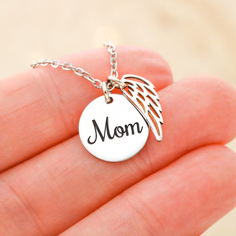 Silver necklace with 'Mom' pendant and angel wing on a hand against a beige background
