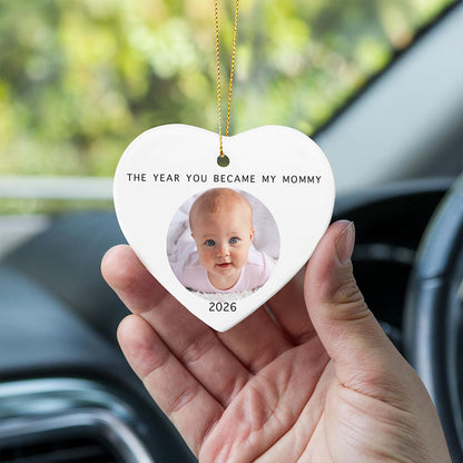 gifts for mom first baby: a hand holds a white heart-shaped ceramic ornament inside a car, featuring a printed photo of a baby, the text 'THE YEAR YOU BECAME MY MOMMY' and '2026', with a gold hanging string.