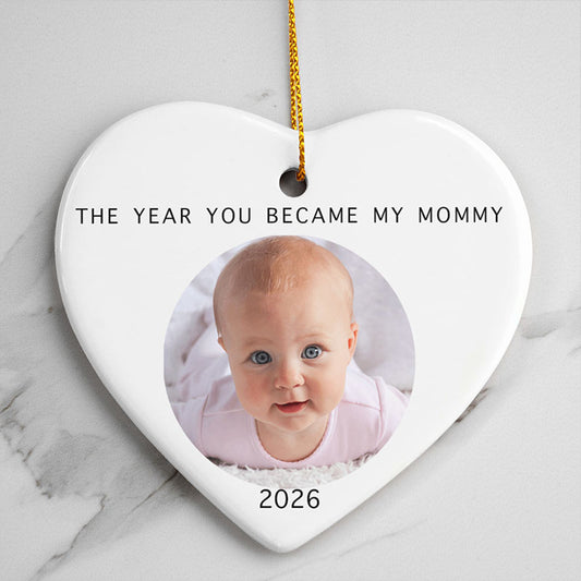 new mom christmas gifts: a white heart-shaped ceramic ornament featuring a baby's photo in the center, printed with text reading "THE YEAR YOU BECAME MY MOMMY" above and "2026" below, hanging from a gold string.