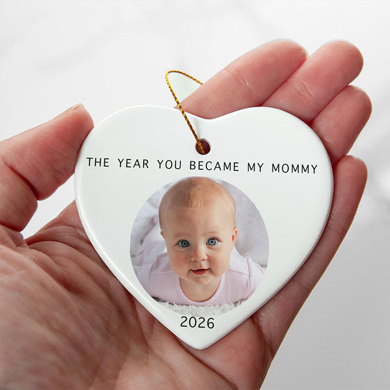 1st time mom gift: A hand holds a white heart-shaped ceramic ornament with a printed photo of a baby, the text 'THE YEAR YOU BECOME MY MOMMY' and '2026', and a gold hanging string against a soft light background.